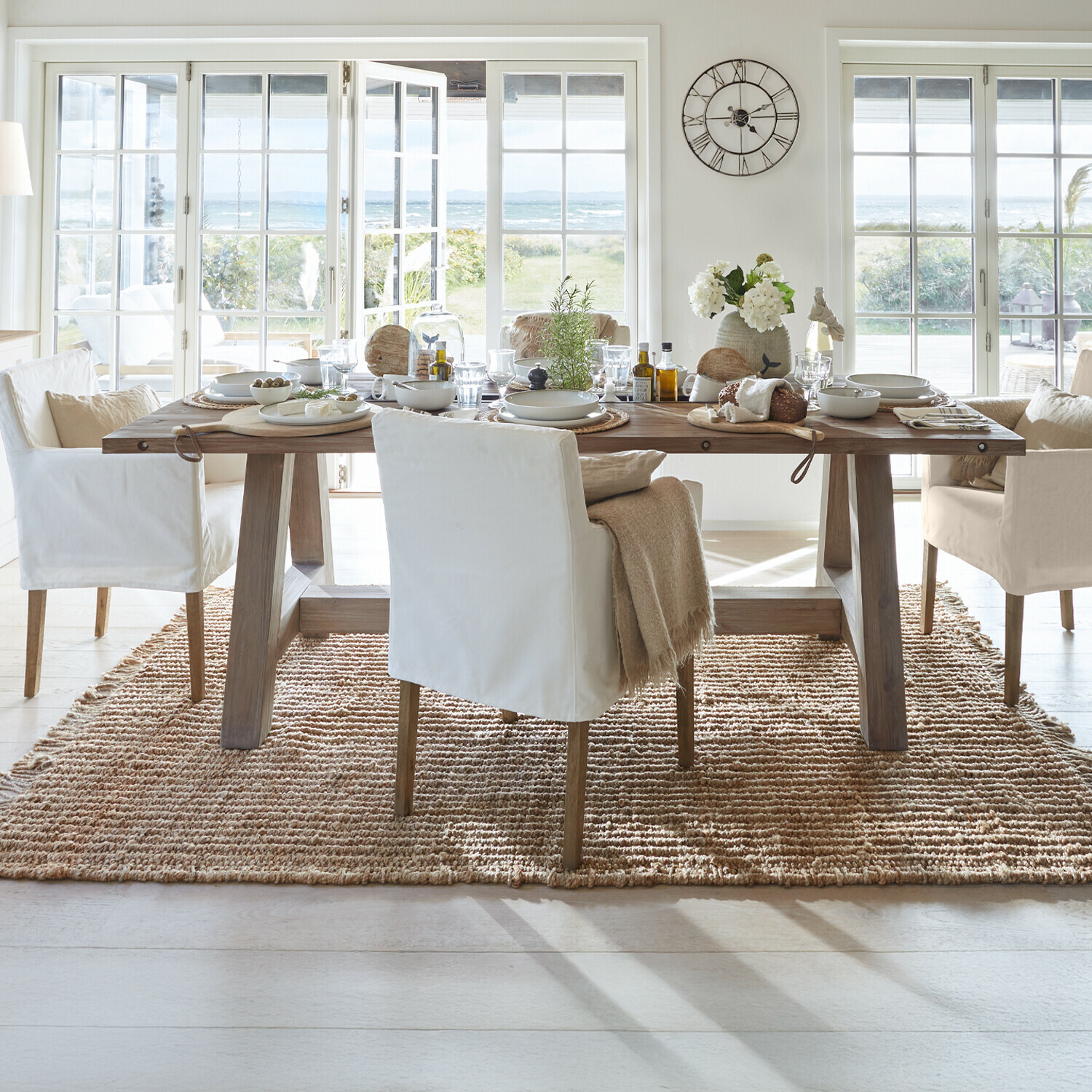 Mesa Berrien Comedor con mesa de madera y sillas blancas sobre alfombra natural con vistas al jardín y al mar