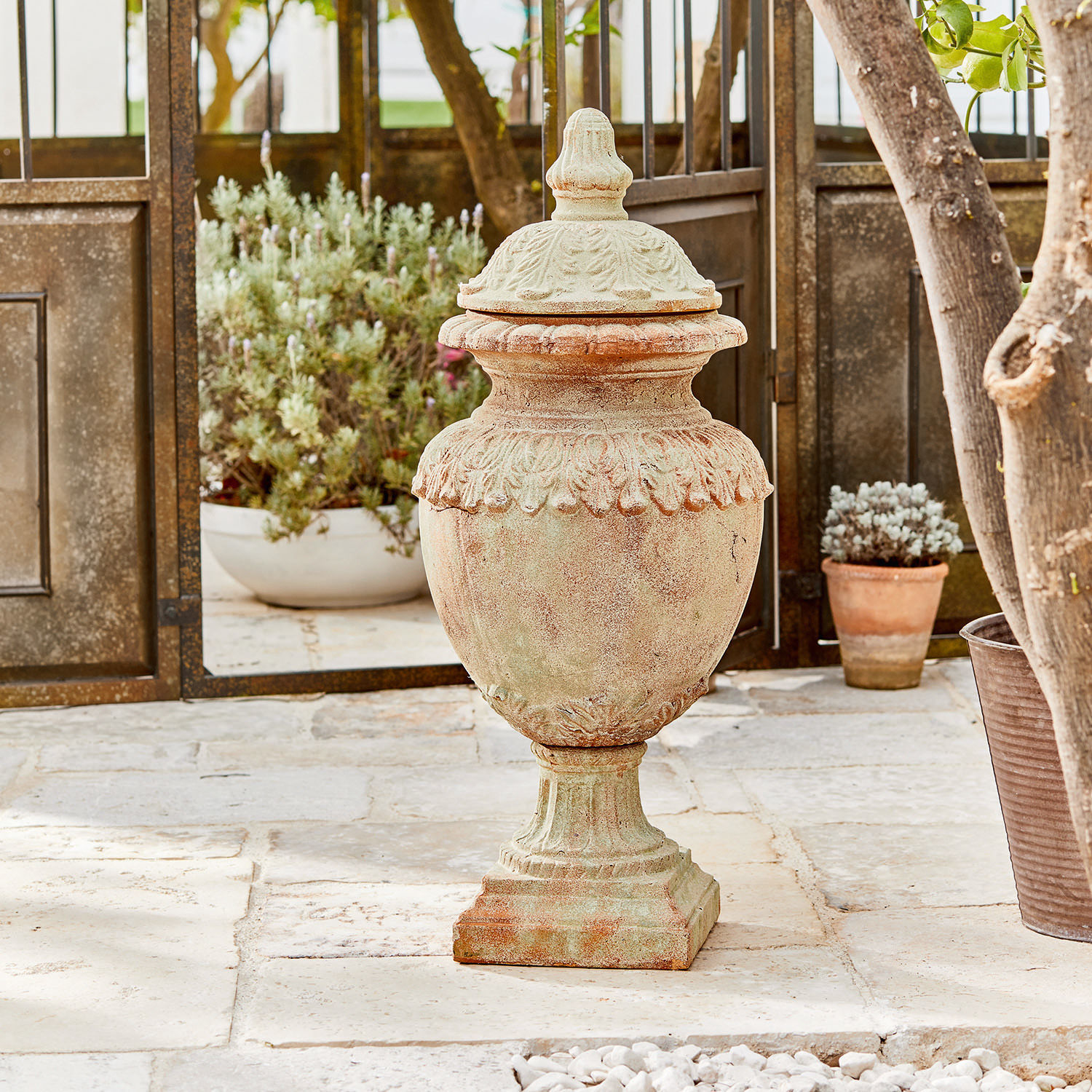 Objeto decorativo Persephone Jarrón decorativo de terracota color natural sobre suelo de piedra con plantas al fondo