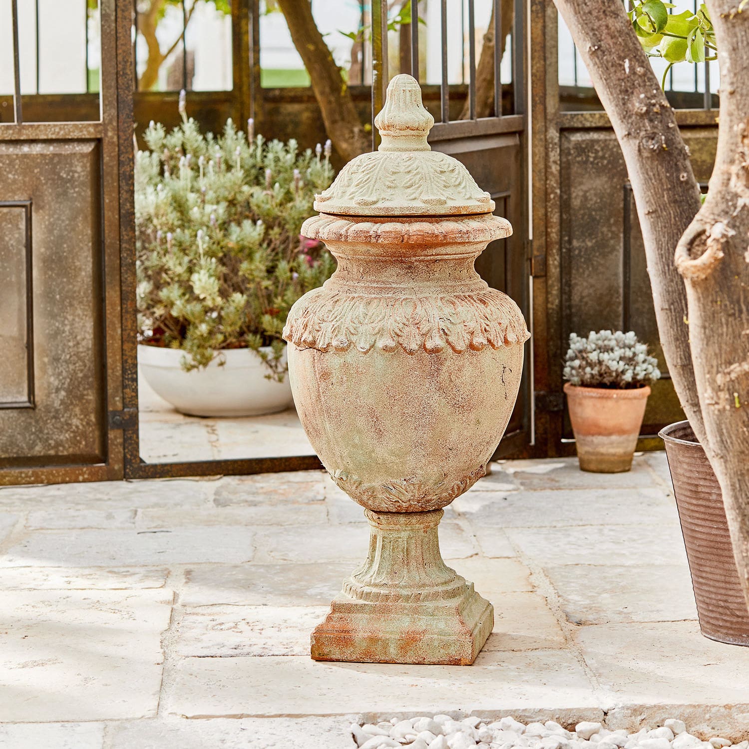 Objeto decorativo Persephone Jarrón decorativo de terracota color natural sobre suelo de piedra con plantas al fondo