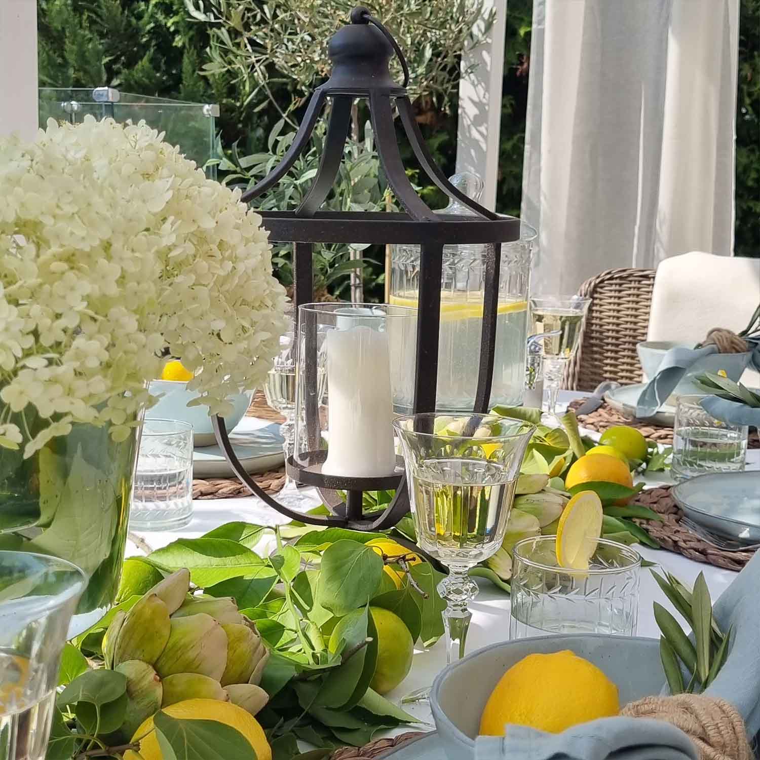 Farol de metal negro con vela blanca sobre mesa de verano decorada con limones y hojas verdes