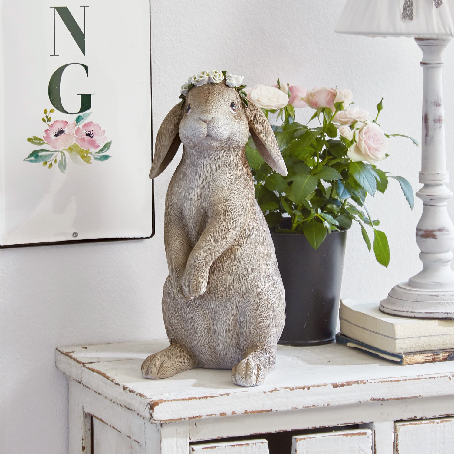 Figura decorativa de conejo beige con corona de flores sobre cómoda blanca de madera