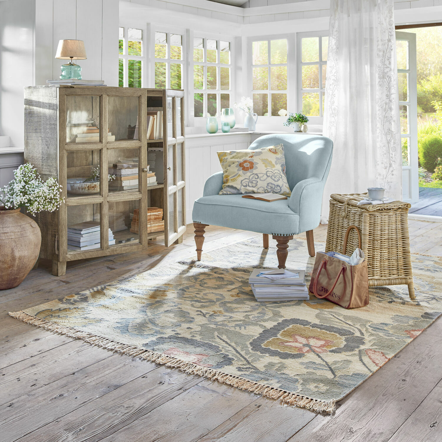 Sala luminosa con sillón azul claro, vitrina de madera y alfombra floral sobre suelo claro