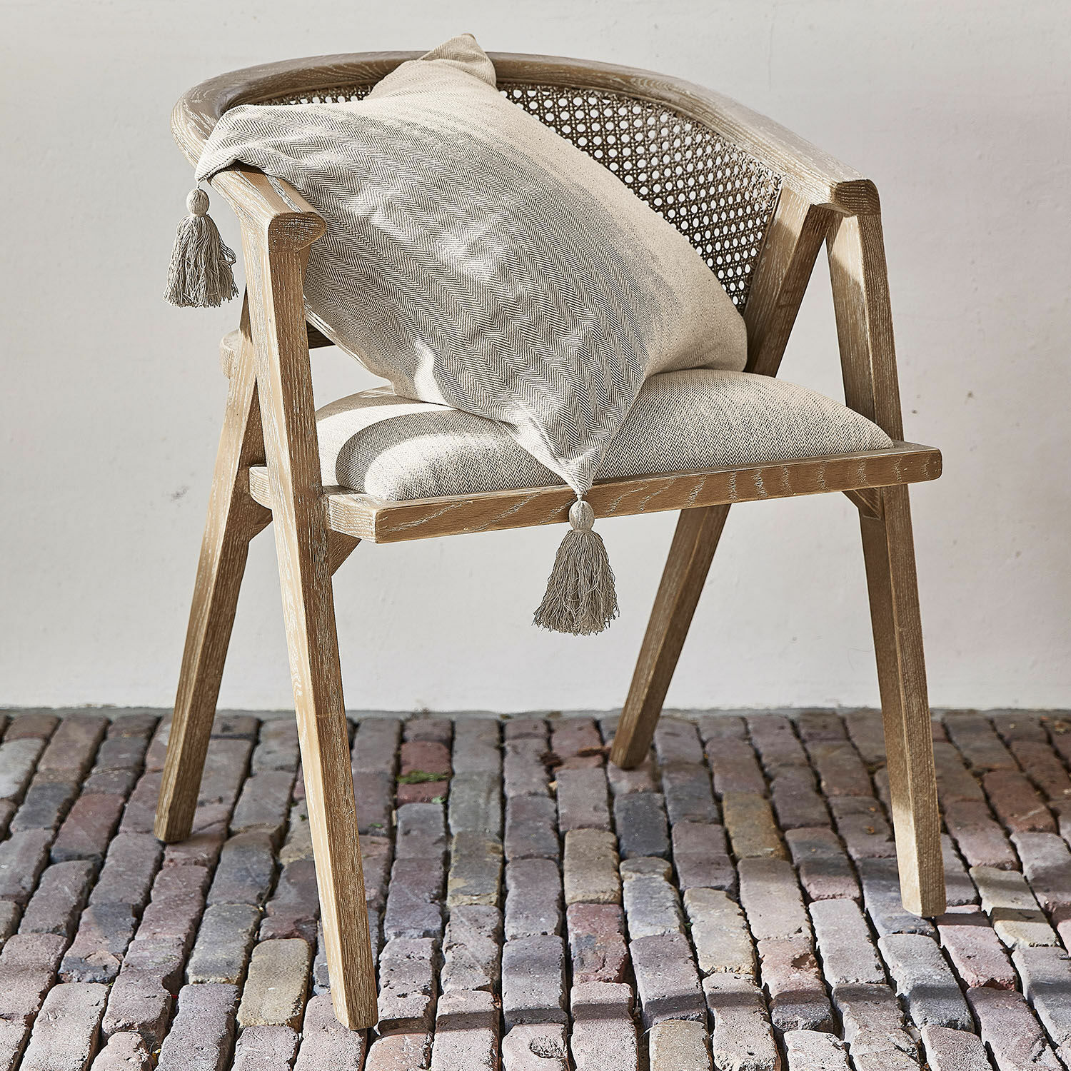 Silla Collaroy Silla de madera clara con respaldo de rejilla y cojín beige con borlas sobre suelo de piedra