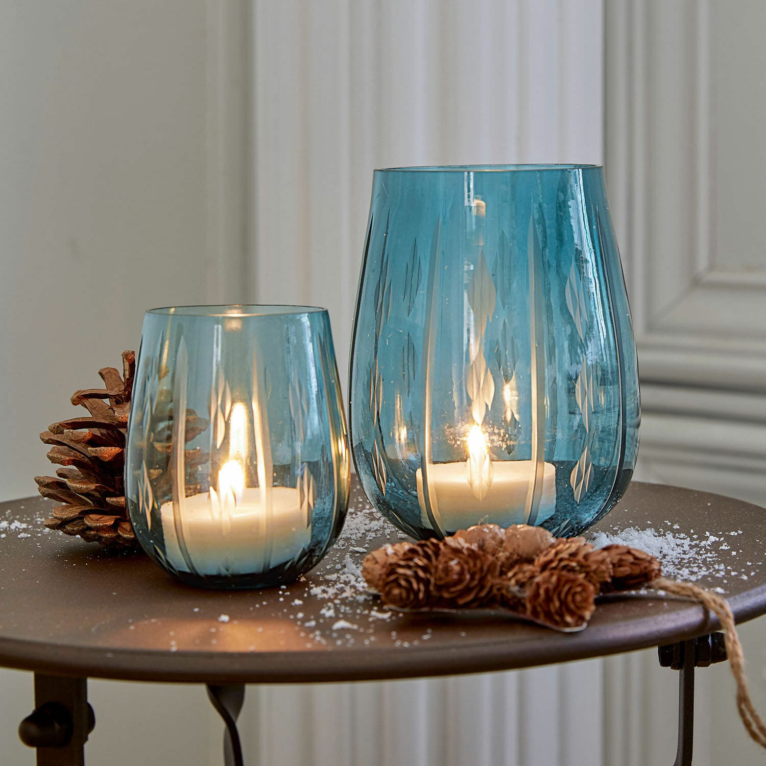 Set de 2 farolillos Danisar Dos portavelas de vidrio azul con patrón, velas blancas encendidas y decoración de piñas en mesa