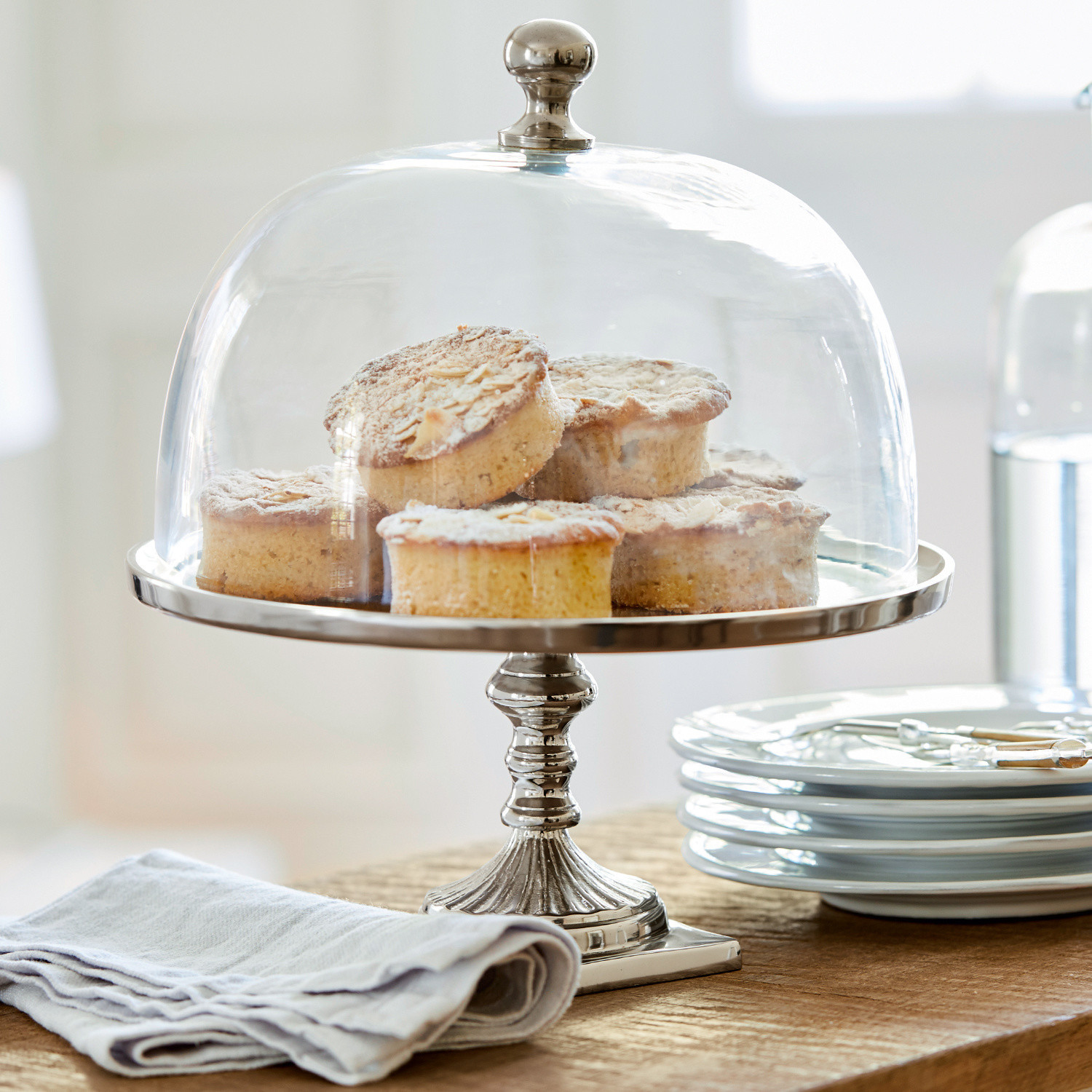 Campana de cristal con plato Mine Plato metálico con campana de vidrio transparente con pasteles sobre mesa de madera con platos blancos