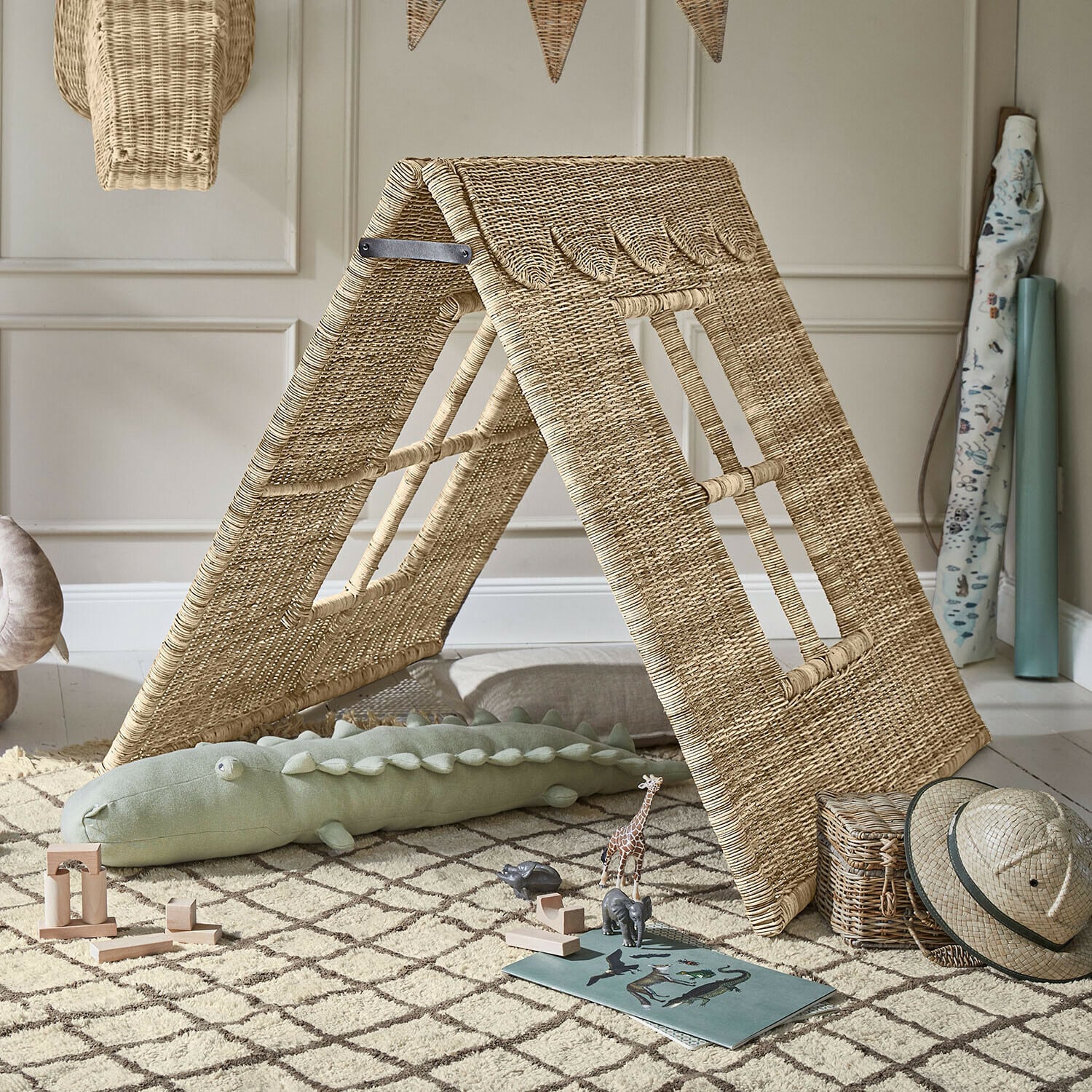 Caseta infantil de ratán natural sobre alfombra con juguetes de madera y sombrero safari