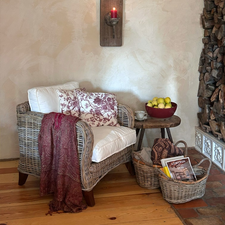 Sillón de mimbre con cojín blanco y manta burdeos junto a mesa de madera con cuenco de frutas y cestas