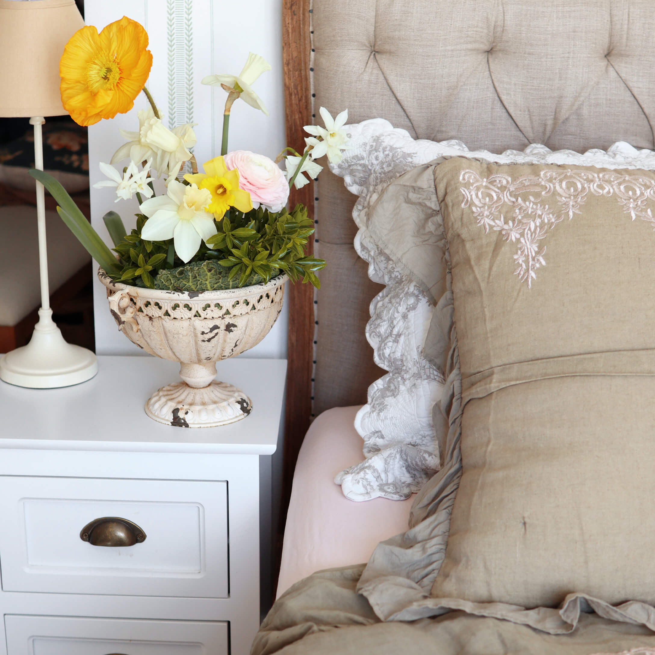 Jarrón decorativo blanco de cerámica con flores de colores sobre mesita blanca junto a la cama