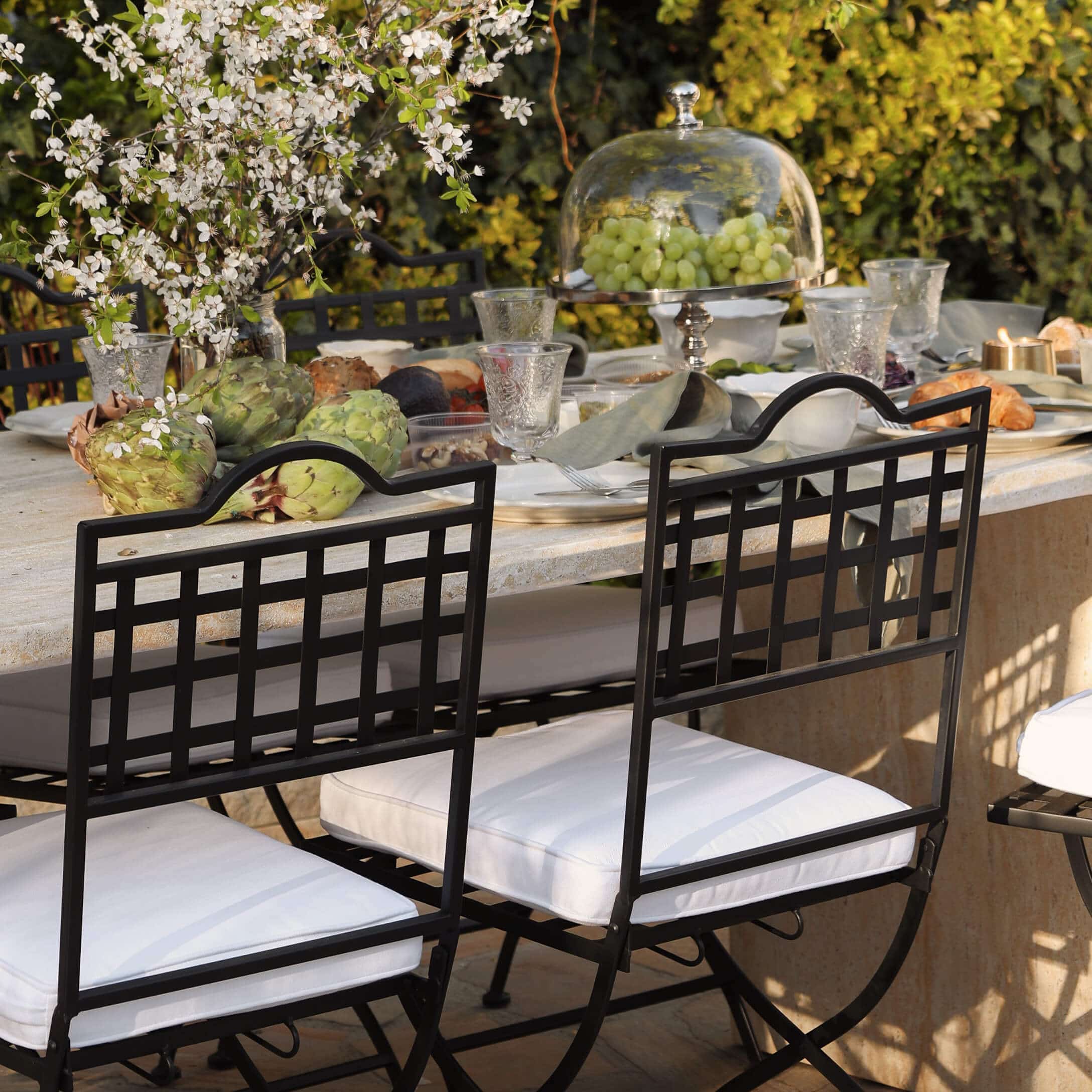 Sillas de metal negro con cojines blancos junto a mesa de piedra con decoración y comida al aire libre