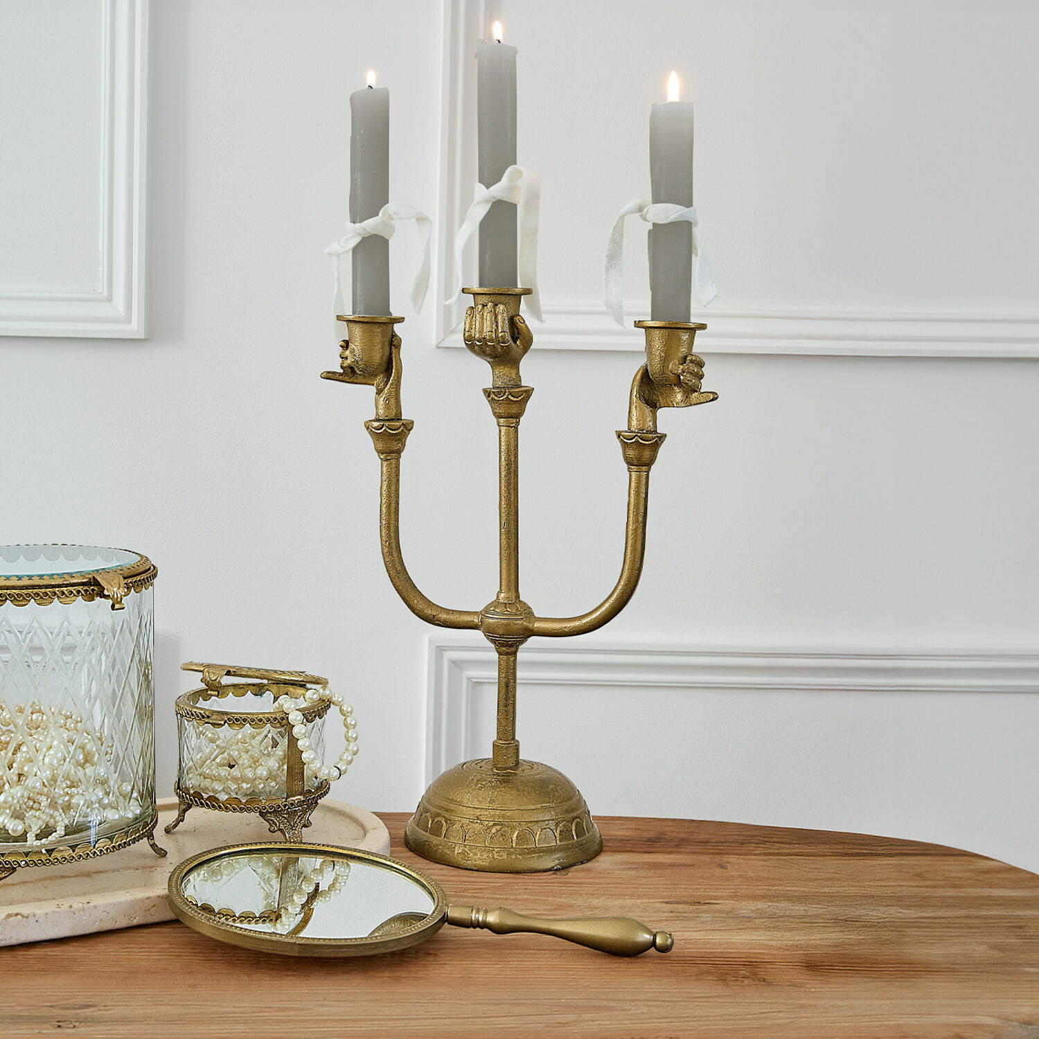 Candelabro dorado con tres velas grises y soportes en forma de manos sobre mesa de madera