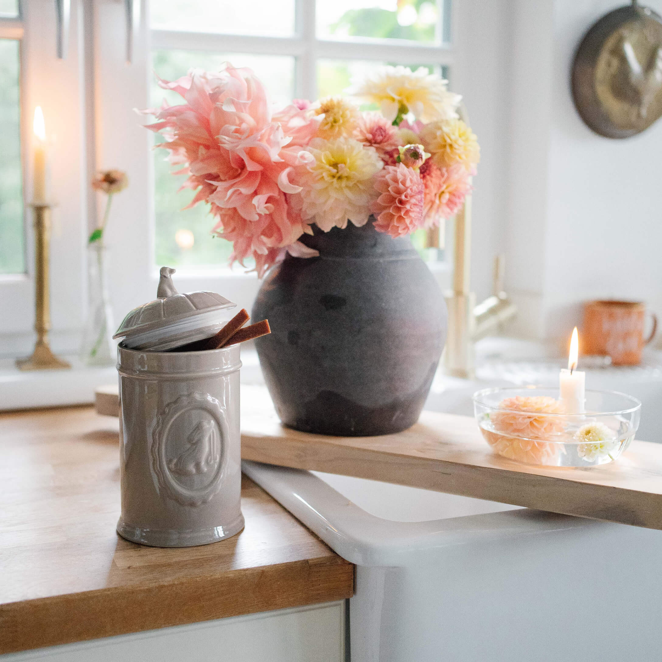 Tarro de cerámica gris con tapa sobre encimera de madera junto a jarrón negro con flores rosa y amarillas
