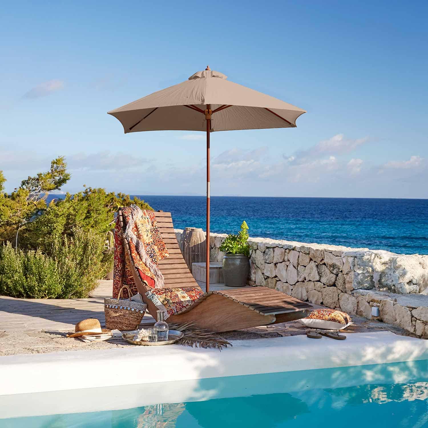 Tumbona de madera con sombrilla beige junto a piscina con vistas al mar y manta colorida