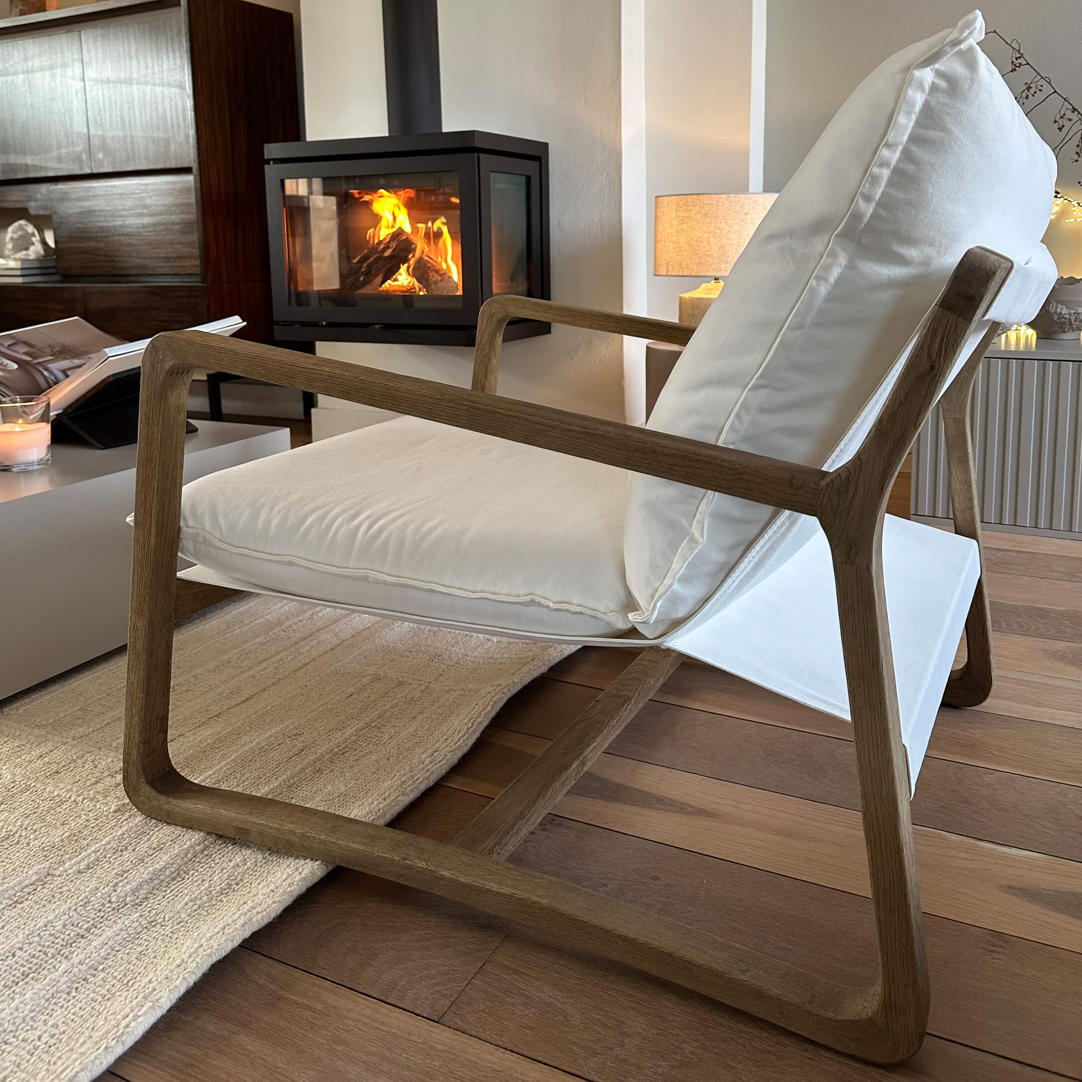 Sillón moderno con estructura de madera y tela blanca en salón acogedor con chimenea encendida