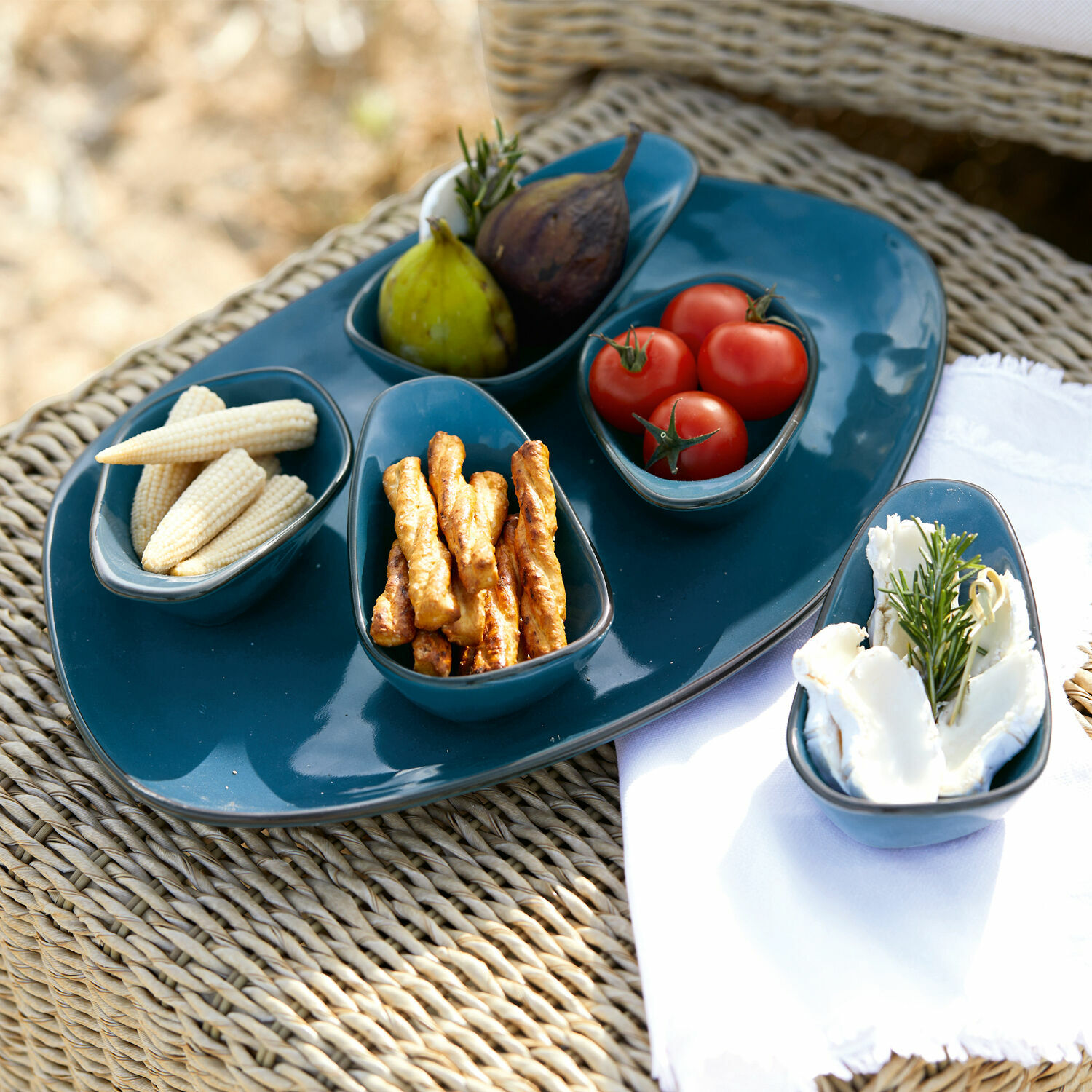 Set de 5 cuencos y plato de servir Biarré Conjunto de cuencos y plato de cerámica azul con aperitivos sobre mesa de mimbre