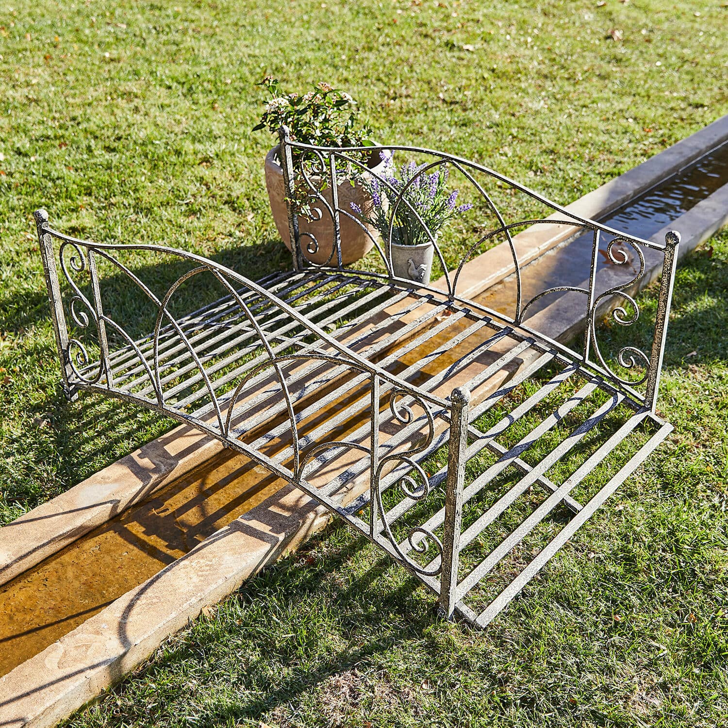 Puente decorativo Escàs Pequeño puente de jardín de metal envejecido en plata con detalles decorativos sobre césped y agua