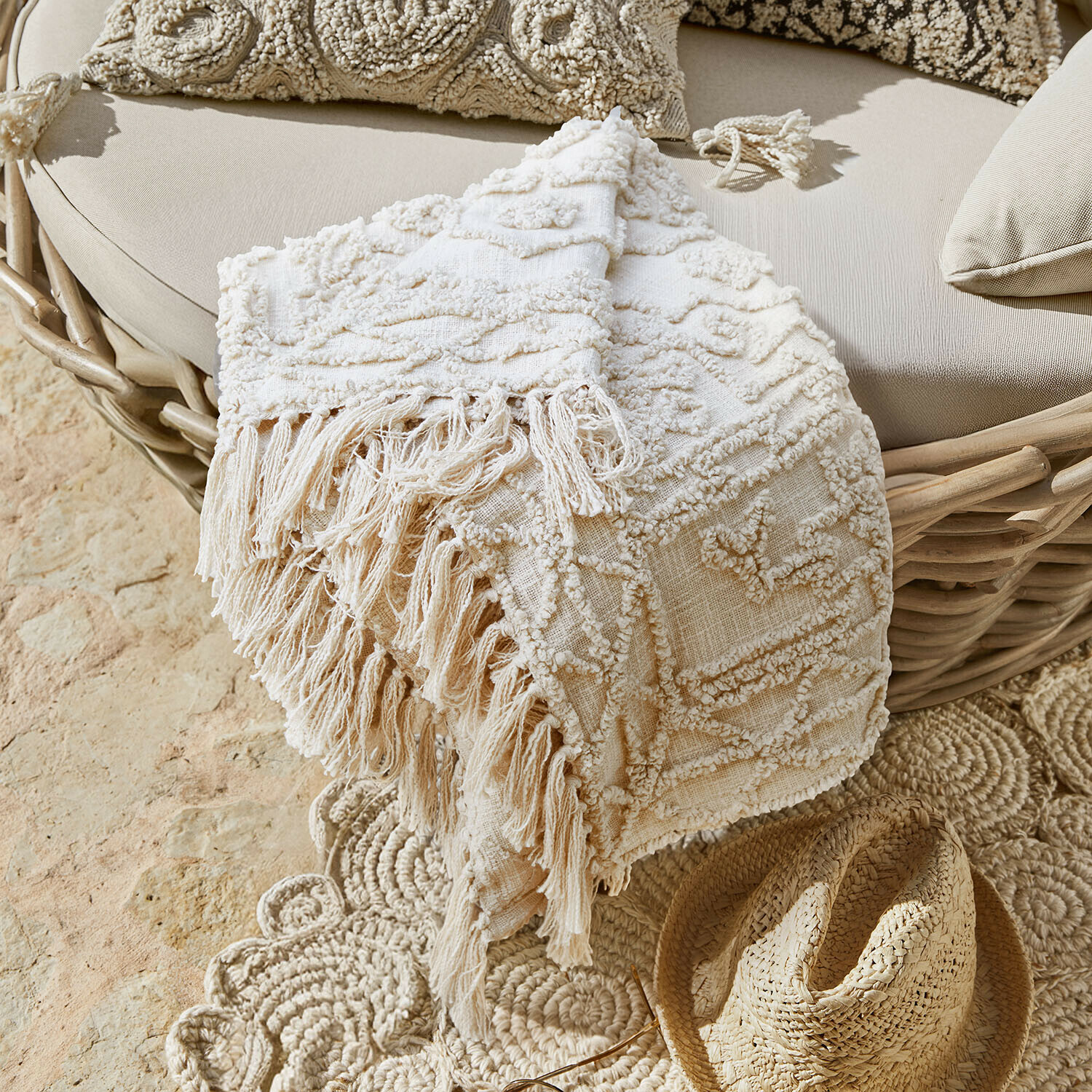 Plaid Norlinn Manta decorativa beige con flecos sobre sillón de mimbre y sombrero de paja en el suelo