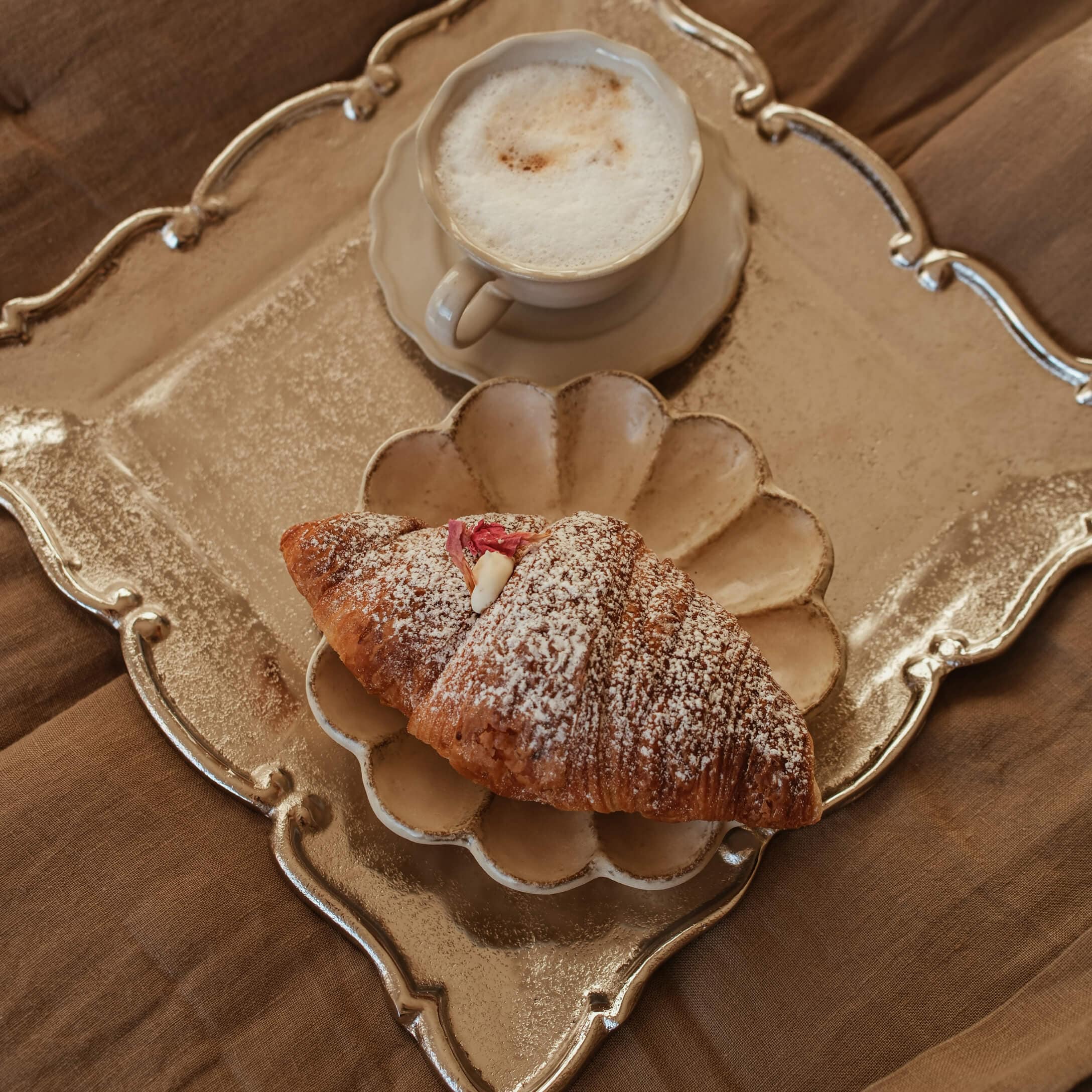 Taza de cappuccino cremoso y croissant con azúcar glas en plato beige y bandeja dorada
