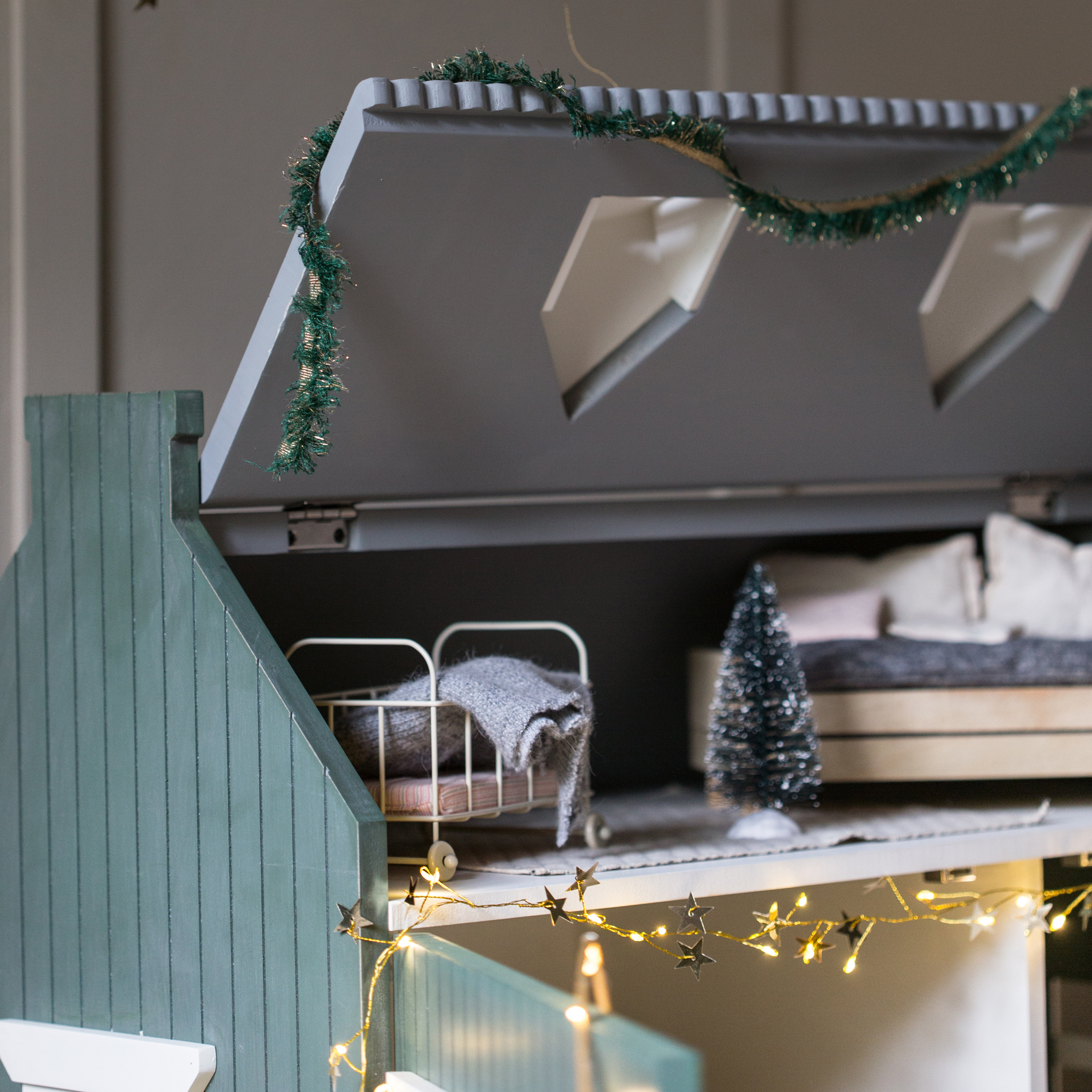 Casa de muñecas de madera con techo verde, cama pequeña y guirnalda de luces decorativa