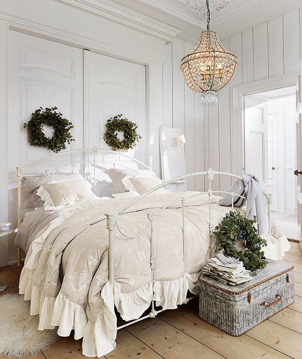 Dormitorio acogedor con cama metálica blanca y ropa de cama beige con motivos florales, coronas verdes decorativas