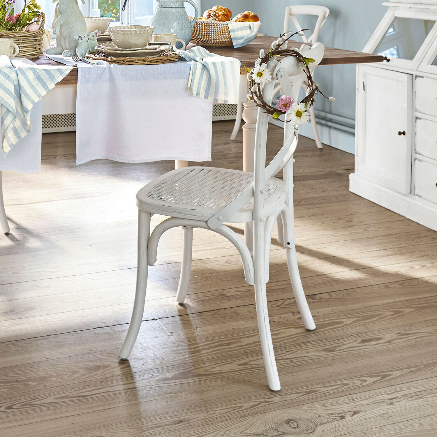 Silla de madera blanca con asiento de mimbre y corona floral decorativa en comedor luminoso