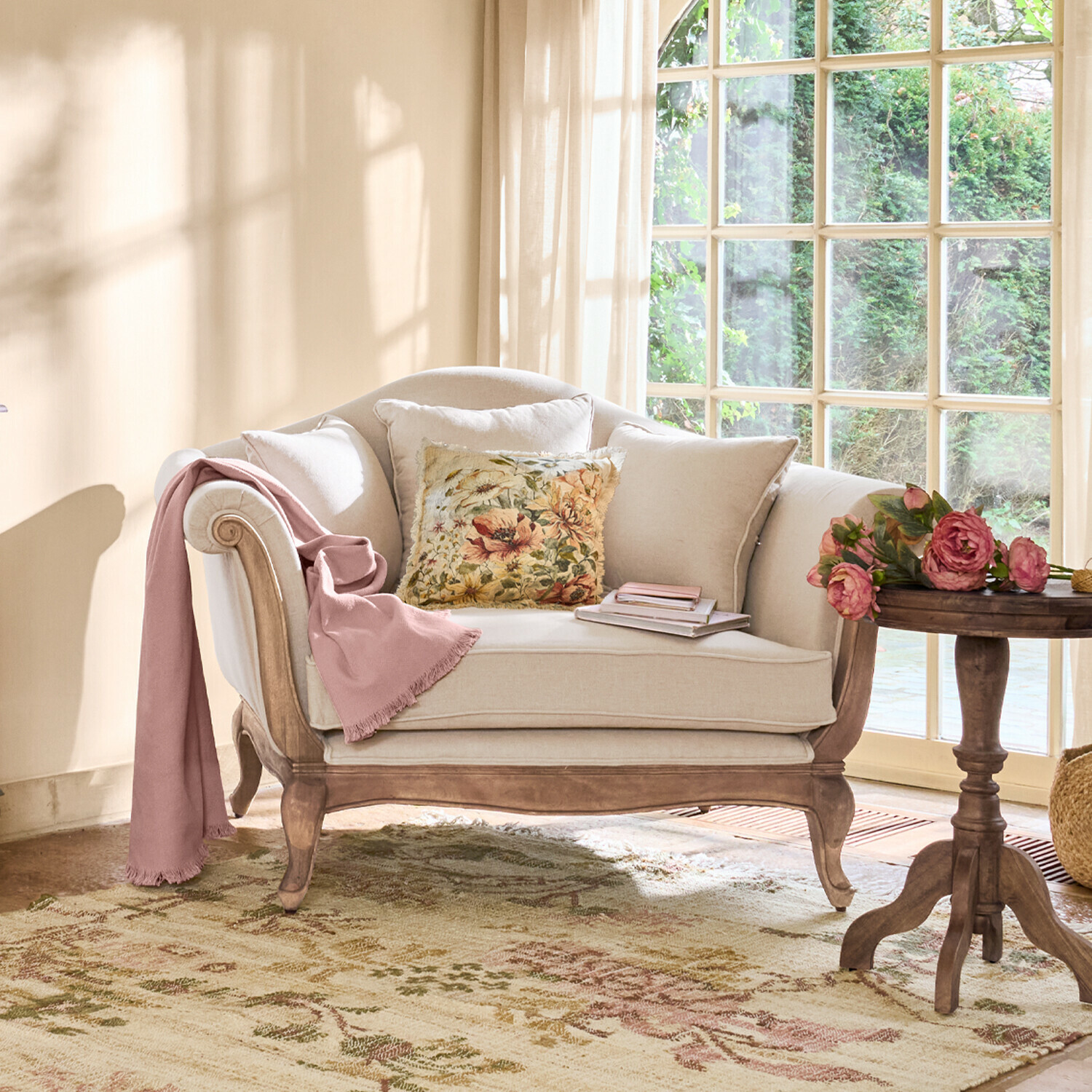 Sofá beige con estructura de madera, cojín floral y manta rosa en salón luminoso con ventana grande