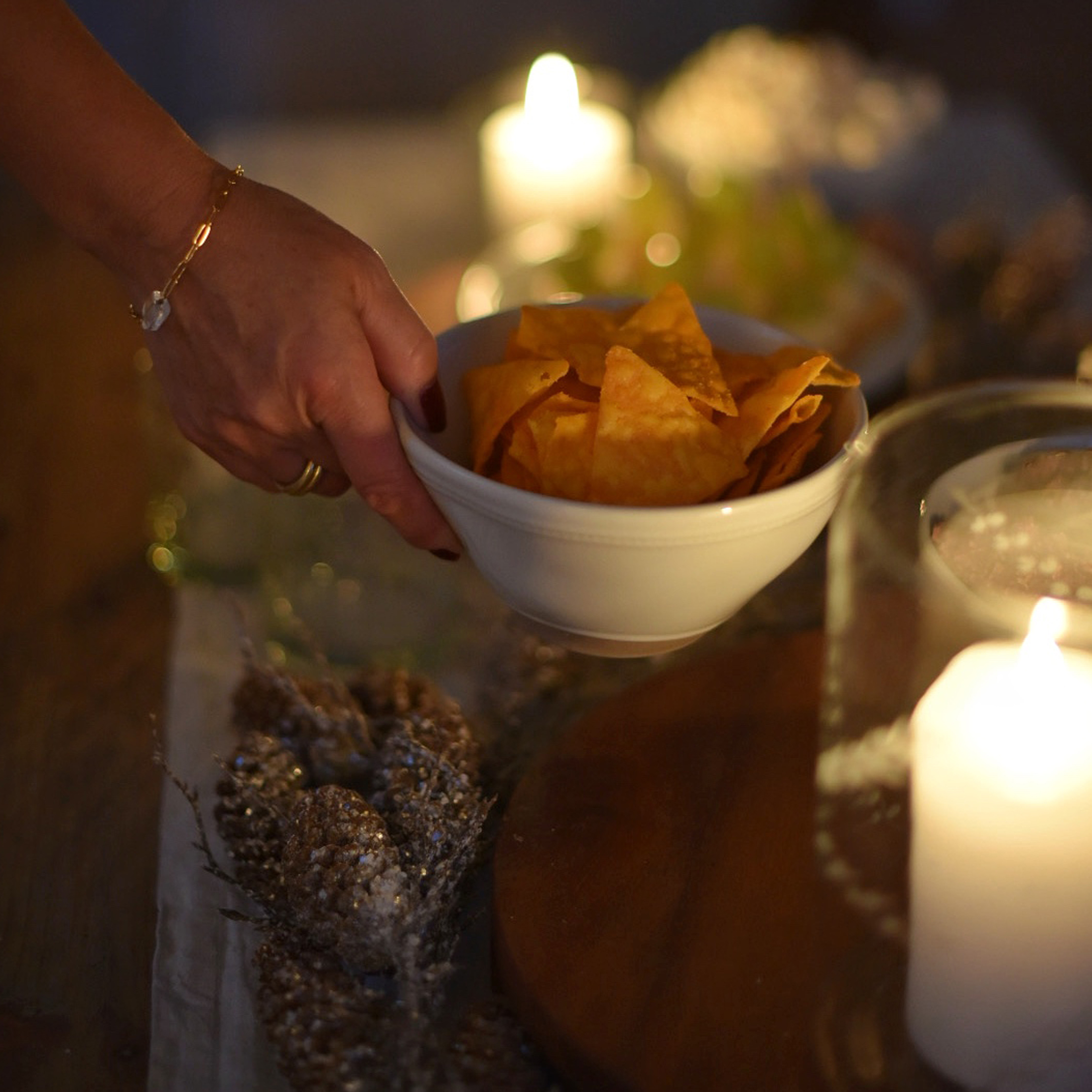 Mano sosteniendo un bol blanco con chips naranjas frente a velas y decoración festiva desenfocada