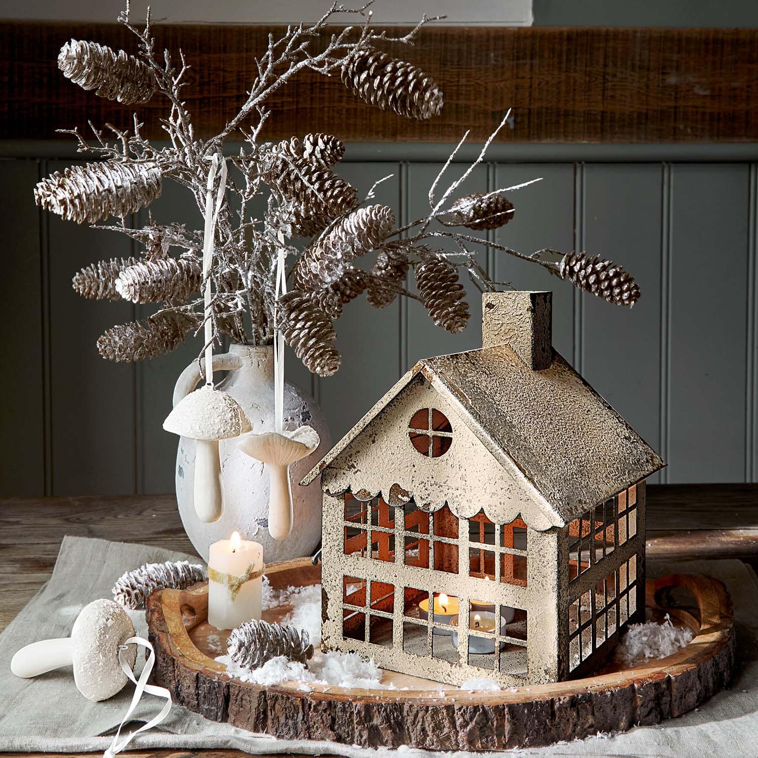 Farol decorativo de metal beige en forma de casa con ramas de piñas y vela sobre bandeja de madera