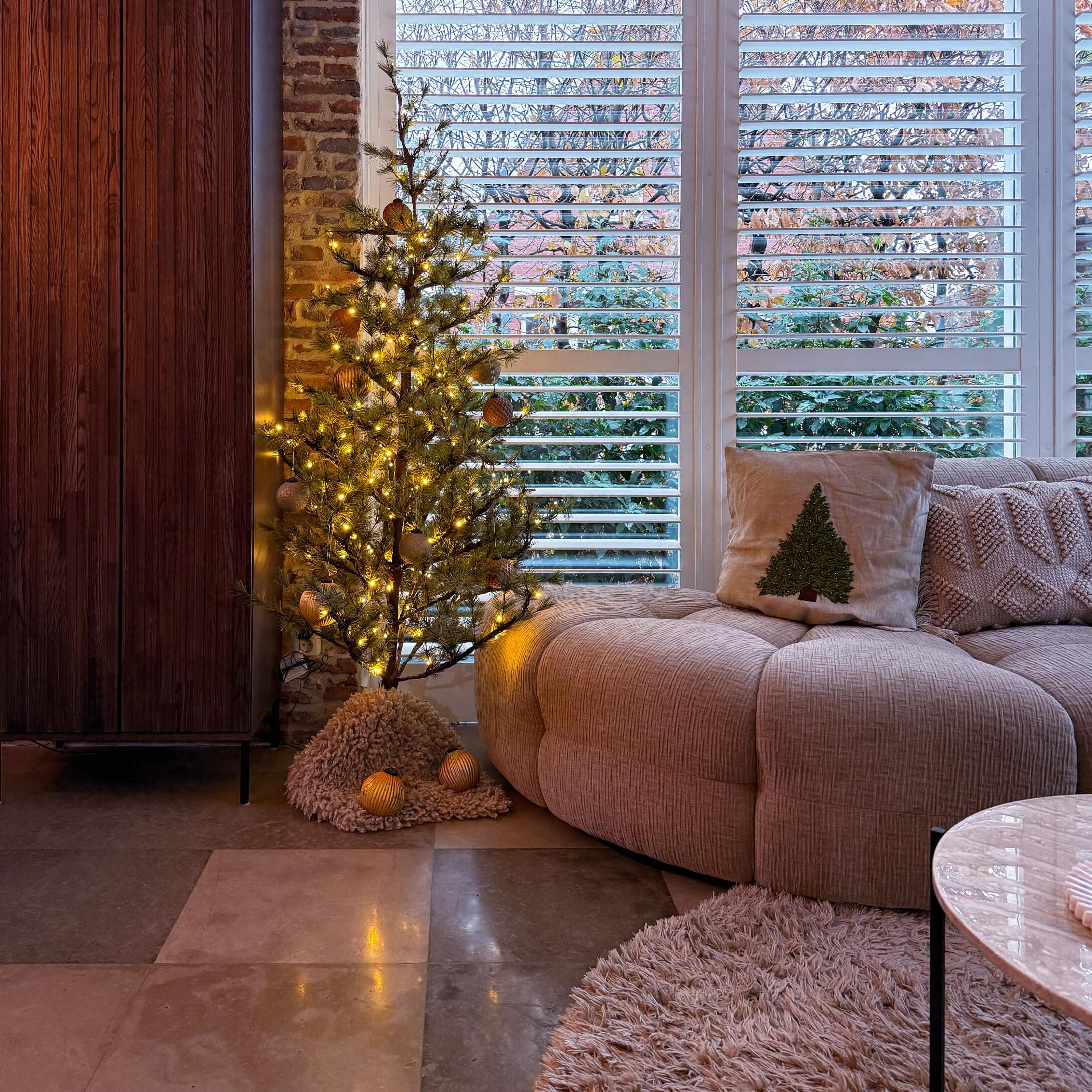 Sala acogedora con sofá beige, cojín con árbol de Navidad y pequeño árbol decorativo iluminado con LED
