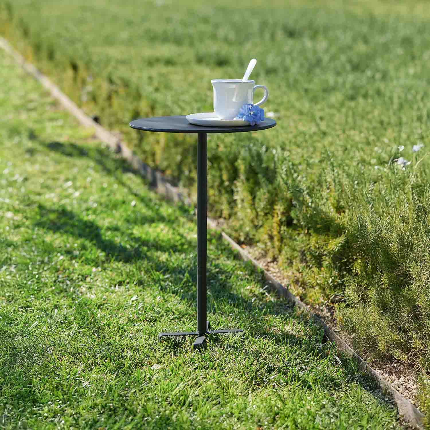 Pequeña mesa de jardín negra de metal con taza blanca y flor azul sobre césped