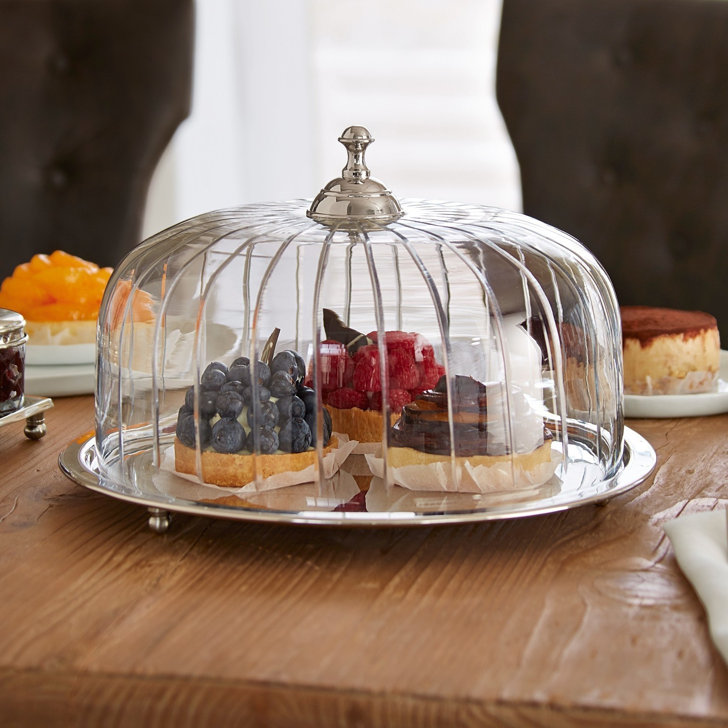 Campana para tarta Allaire Campana de cristal con asa metálica sobre mesa de madera con tartas de frutas variadas