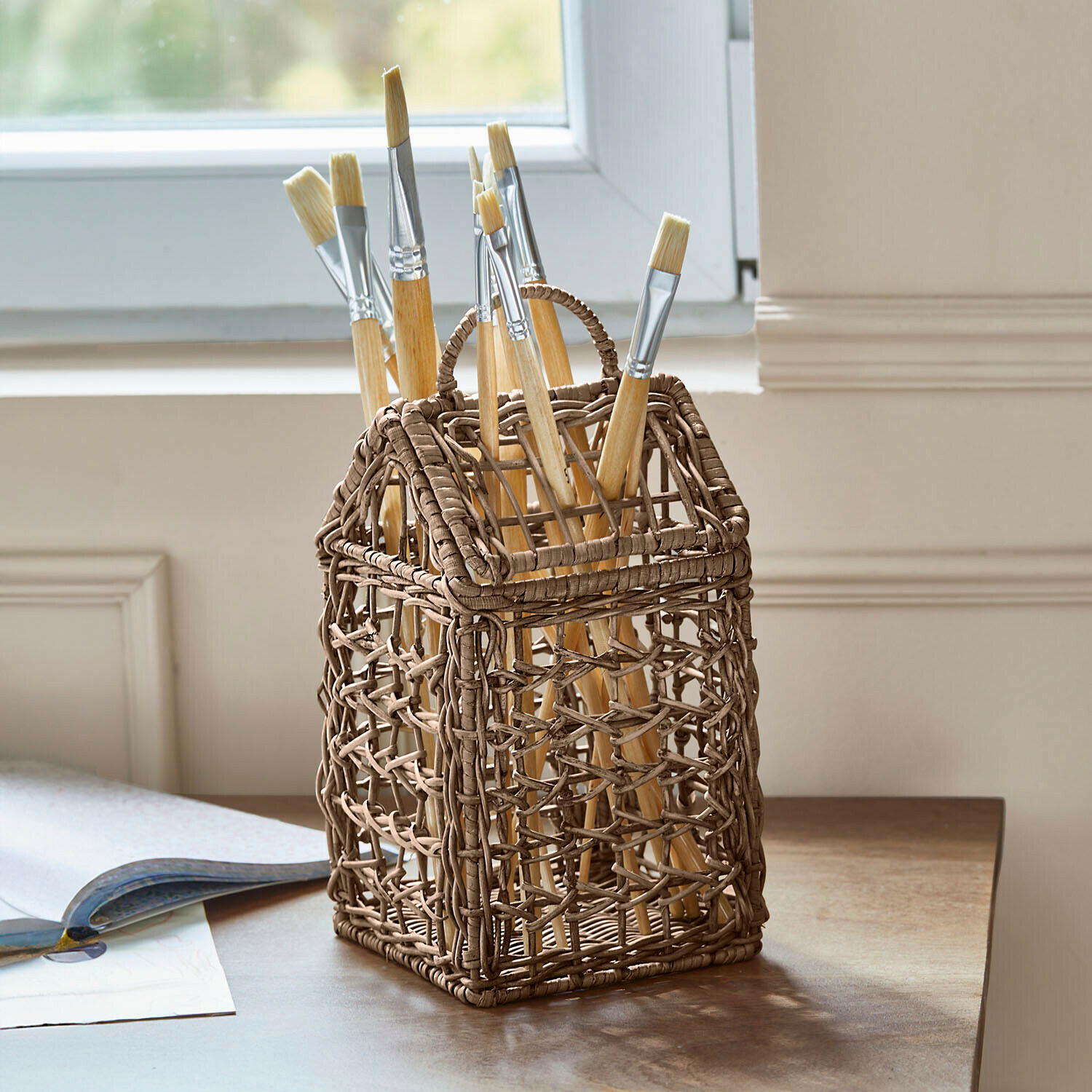 Contenedor de mimbre marrón en forma de casa con pinceles de pintura sobre mesa de madera