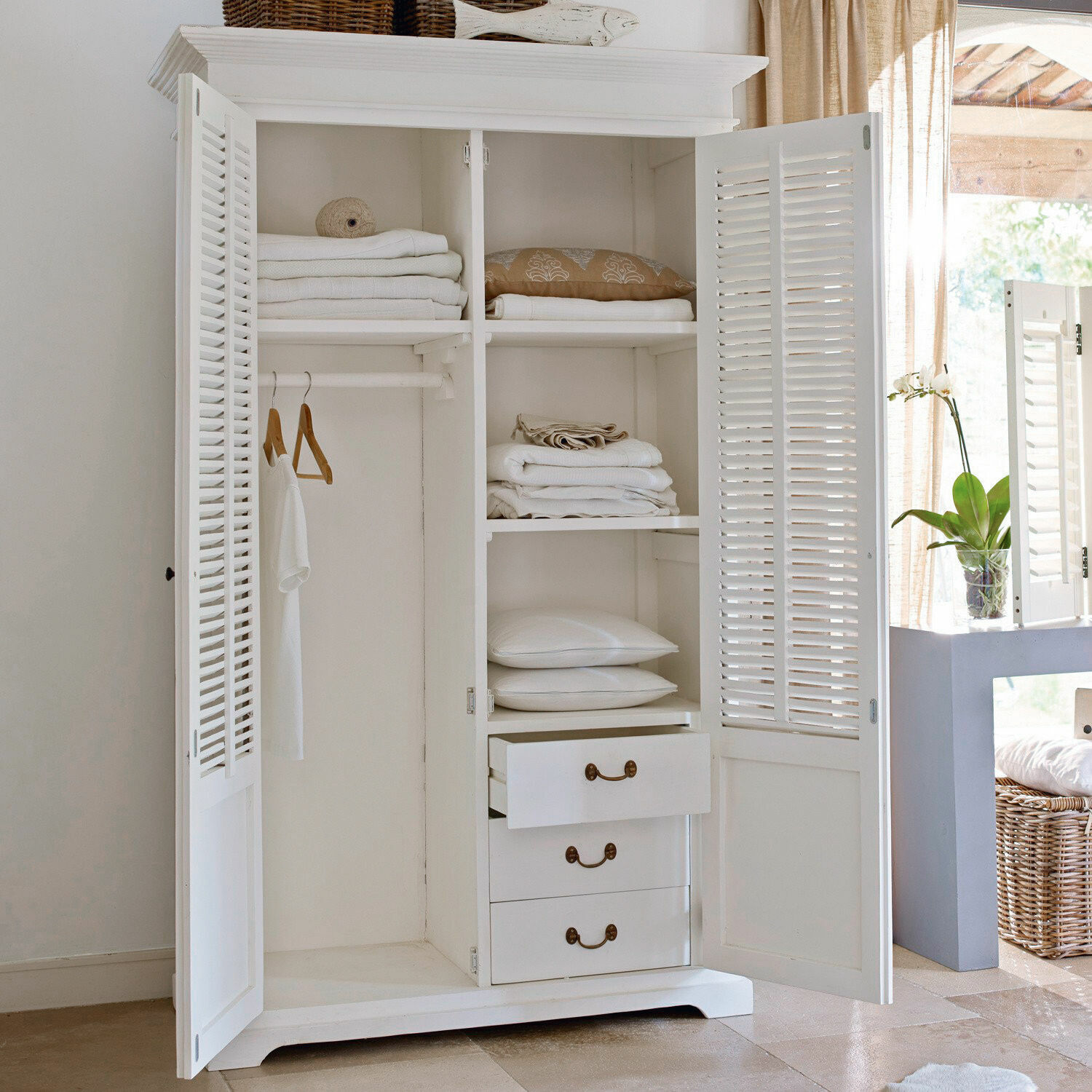 Armario de madera blanco con puertas de persiana y estantes para ropa de cama en habitación luminosa