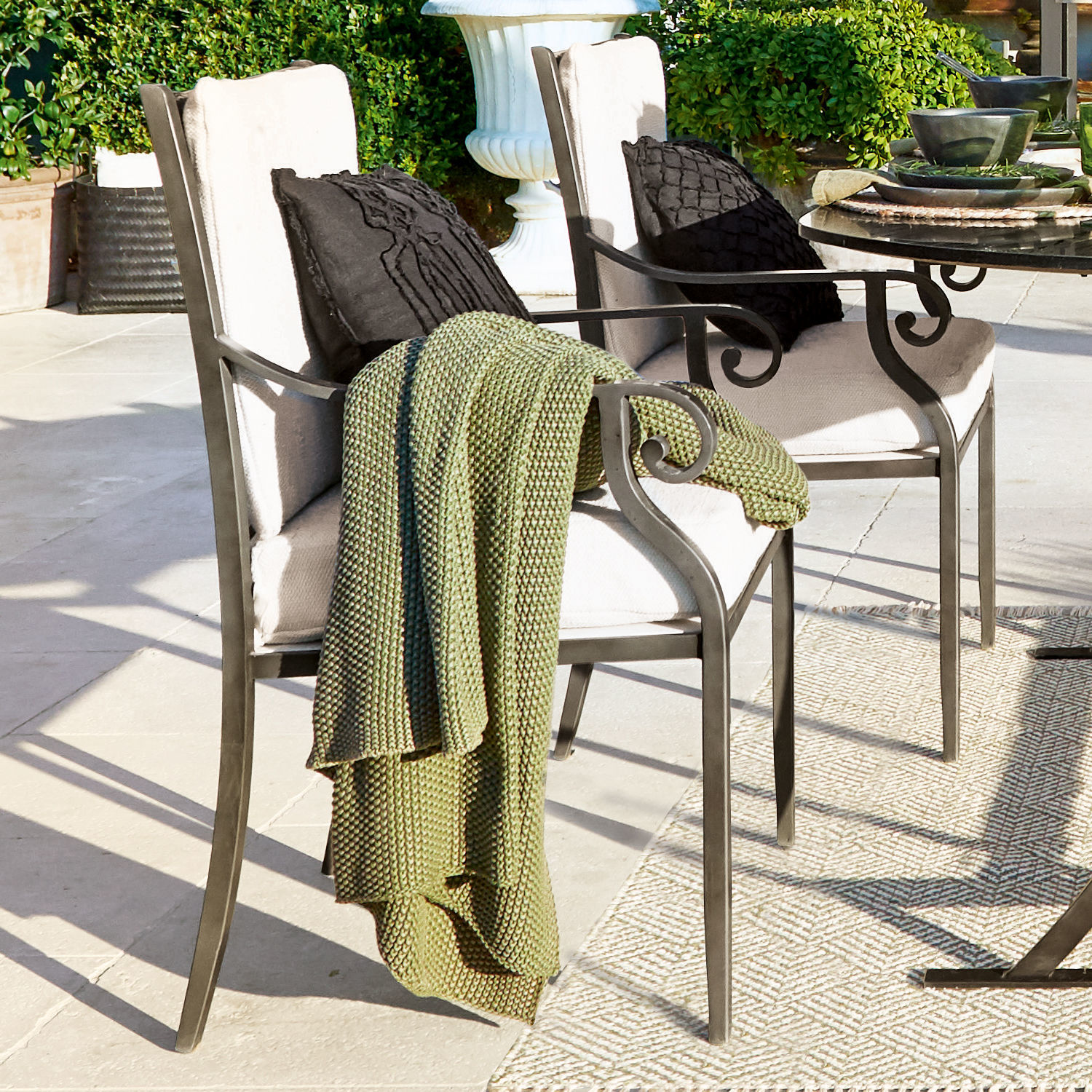 Sillas de jardín de metal con cojines crema y manta verde tejida en terraza de piedra clara