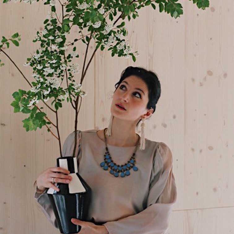 Mujer sostiene jarrón de cerámica blanco y negro con ramas verdes frente a pared de madera clara