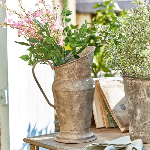 Jarra decorativa de metal envejecido con flores verdes y rosas sobre mesa de jardín iluminada