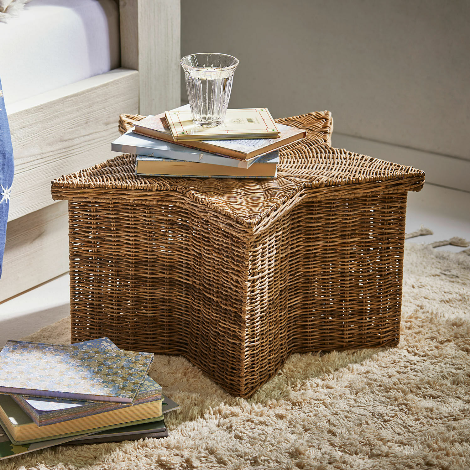 Cesta Rivolina Cesta de ratán en forma de estrella sobre alfombra con libros y vaso de agua, color natural