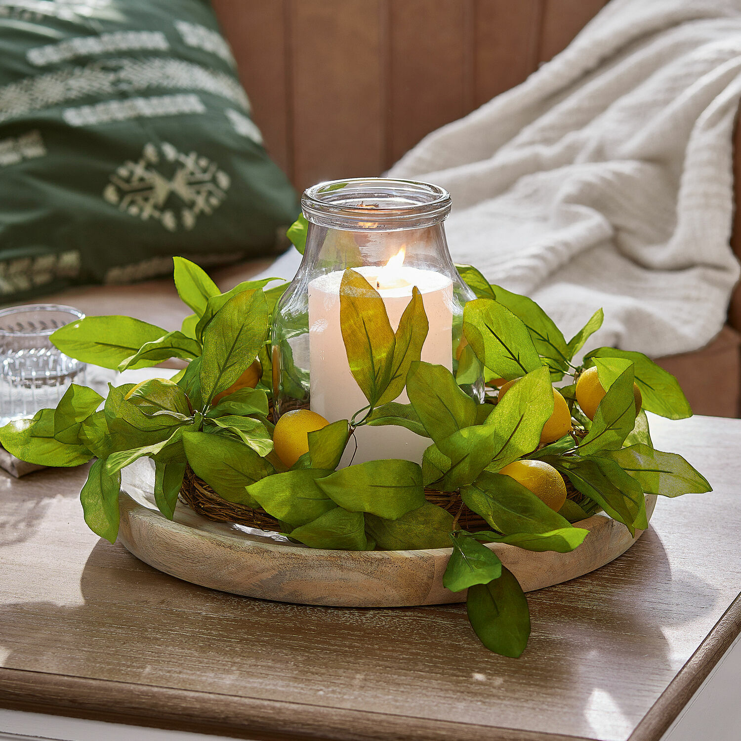 Portavelas de vidrio con vela blanca rodeada de hojas verdes y frutos amarillos sobre bandeja de madera