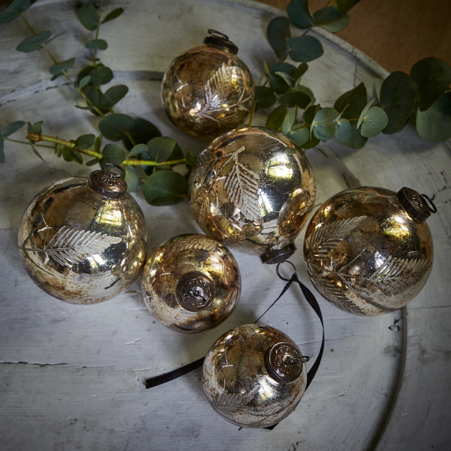 Set de 6 adornos navideños Tristan Conjunto de seis bolas navideñas de vidrio dorado con diseño de hojas sobre bandeja blanca con ramas verdes