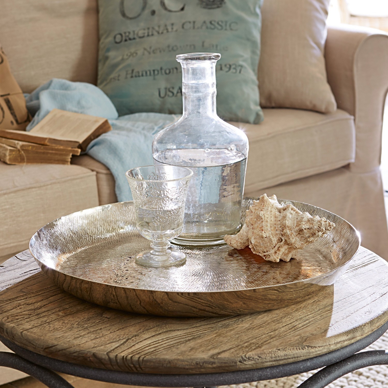 Bandeja Caelan Bandeja redonda de metal plateado sobre mesa de madera con jarra de cristal y concha decorativa