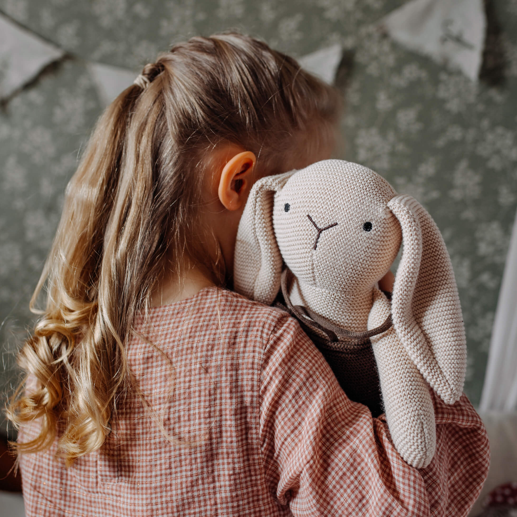 Niña abrazando un conejo de peluche beige tejido en una habitación infantil
