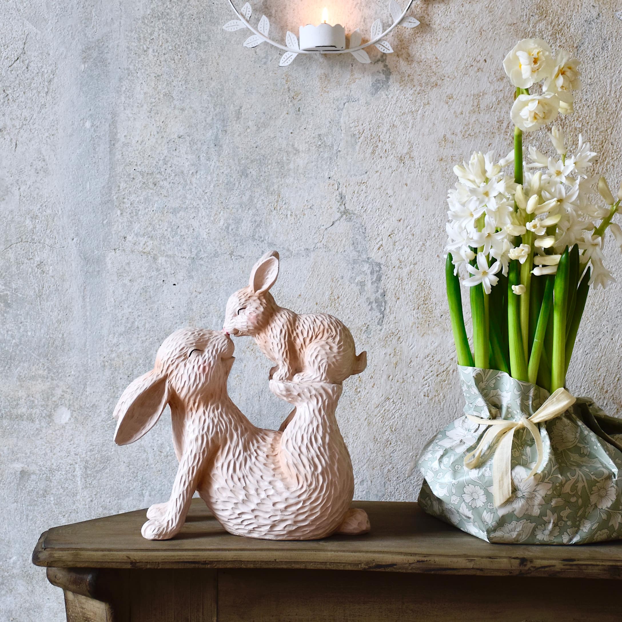 Escultura de madera rosa con dos conejos sobre mesa de madera junto a flores en jarrón pastel