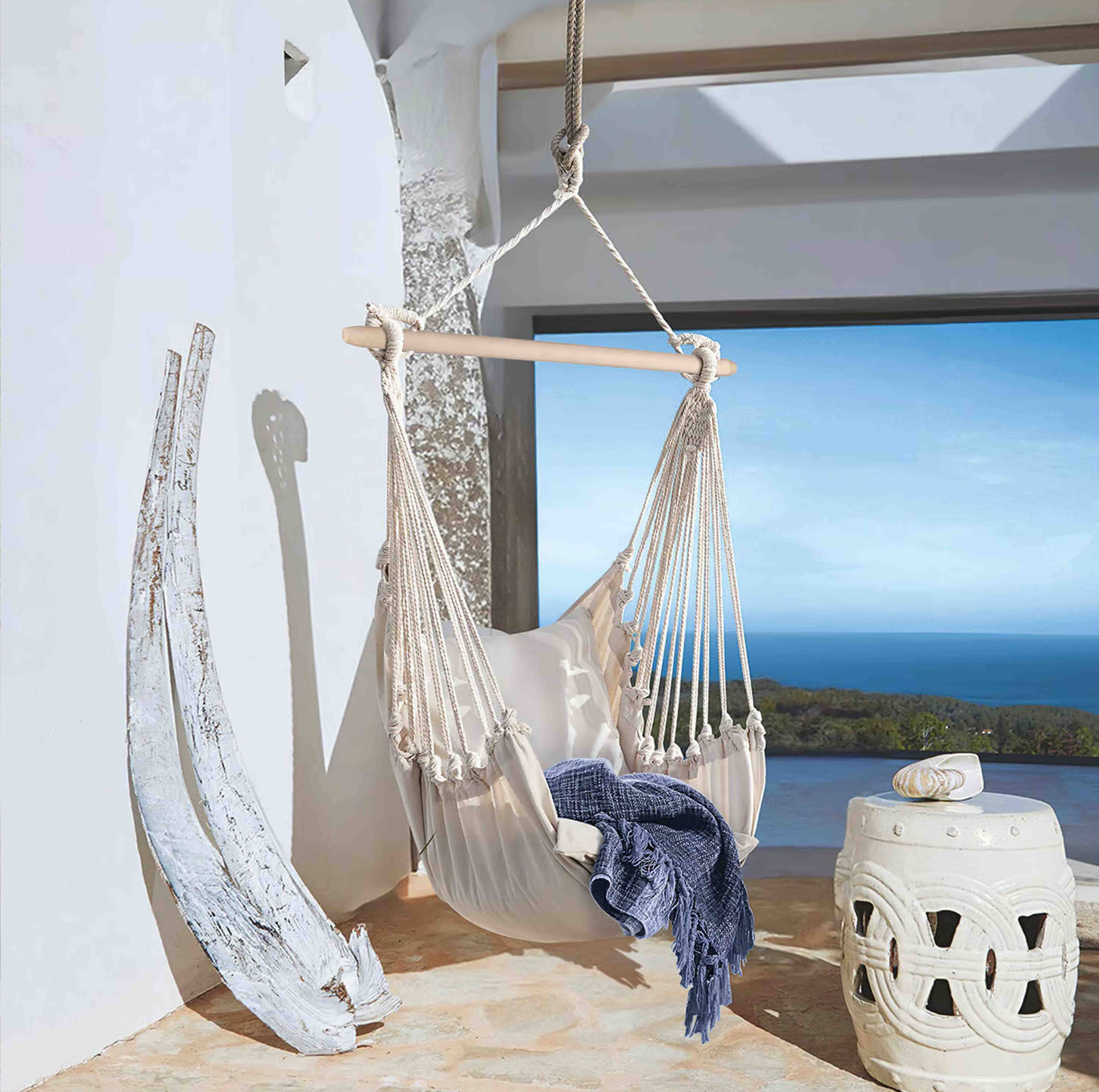 Hamaca beige de algodón con barra de madera y manta azul en terraza con vistas al mar