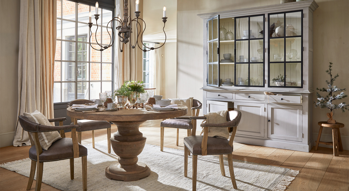 Comedor con mesa redonda de madera y cuatro sillas marrones con reposabrazos, vitrina blanca al fondo