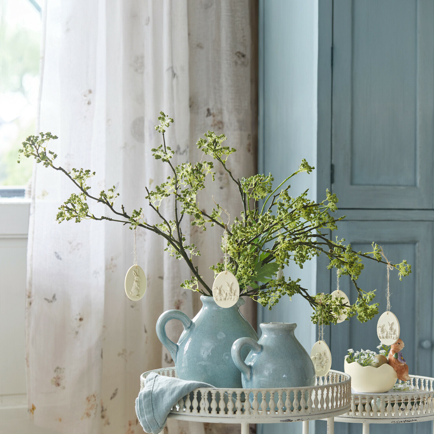 Jarras de cerámica azul con ramas verdes y decoraciones de Pascua sobre mesa metálica blanca