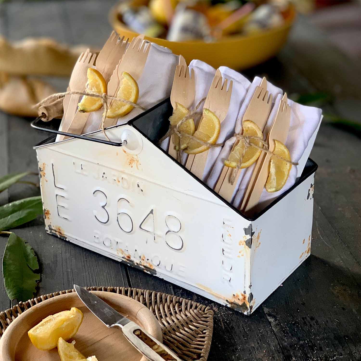 Contenedor metálico blanco envejecido con cubiertos de madera y servilletas blancas decoradas con rodajas de limón