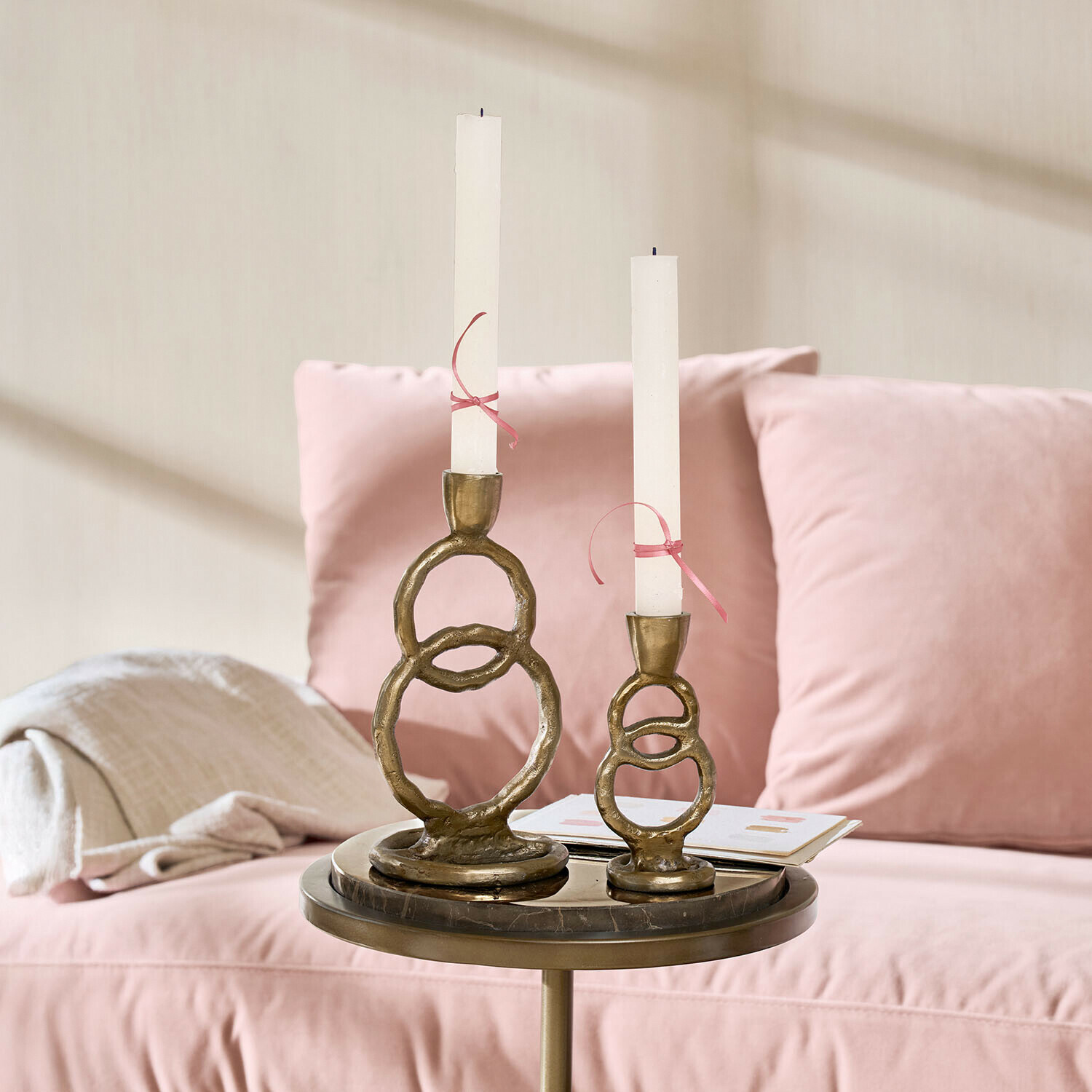 Conjunto de dos candelabros de latón con velas blancas sobre una mesa junto a un sofá rosa