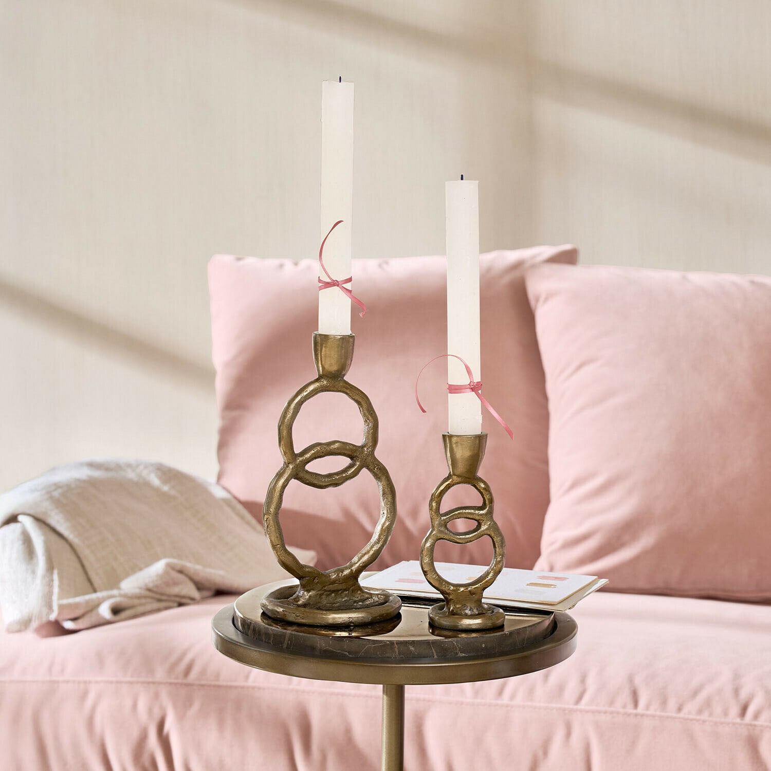 Set de 2 candeleros Corvane Conjunto de dos candelabros de latón con velas blancas sobre una mesa junto a un sofá rosa