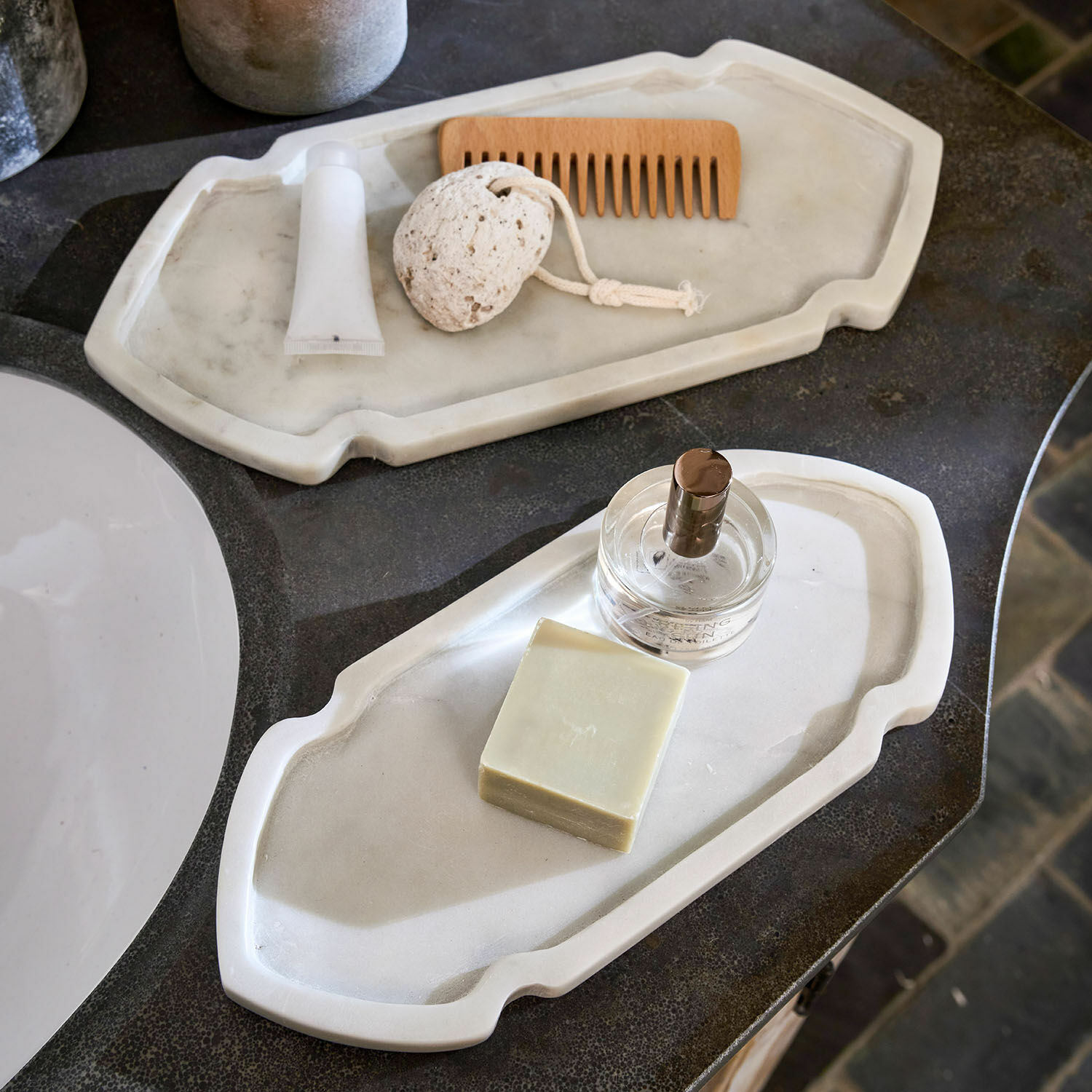 Set de 2 bandejas Zafrina Dos bandejas de mármol blanco con jabón, perfume y peine de madera sobre encimera de baño
