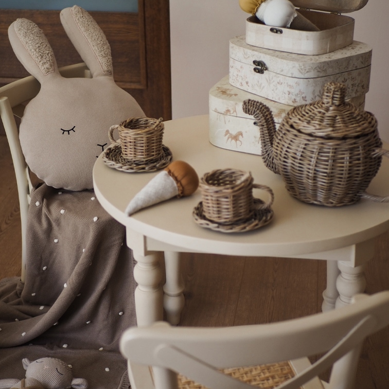 Juego de té de mimbre sobre mesa blanca con cojín de peluche de conejo y manta marrón
