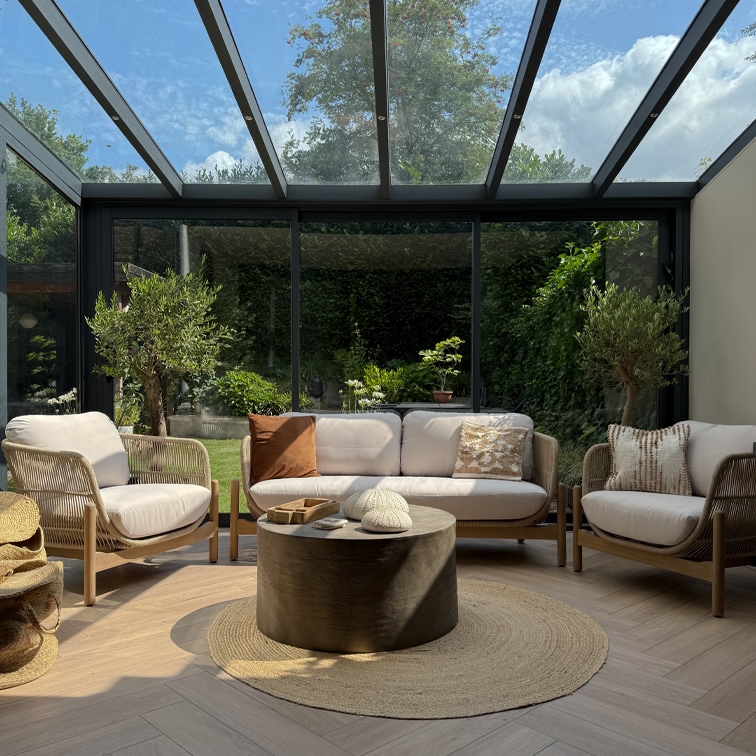 Sala acogedora con sofás y sillones claros de madera y tela bajo techo de cristal, alfombra redonda natural
